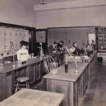 A black and white photo of a woman in a plumed hat in a laboratory classroom with several men