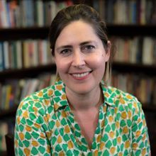 A woman in a colorful blouse smiles in front of a blurred background of books on shelves