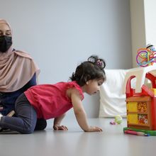 An excited-looking toddler crawls towards toys while her mother watches on.