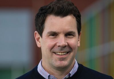 A man wearing a dark blue sweater is smiling and standing in front of a building