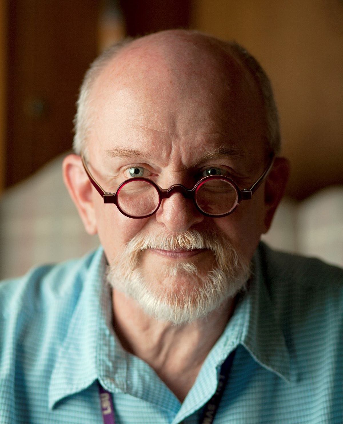 Portrait of man with a white goatee wearing an aquamarine shirt and burgundy spectacles.