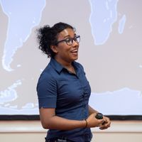 Izzy Jayasinghe stands in front of a projection of a map, looking as though she is speaking