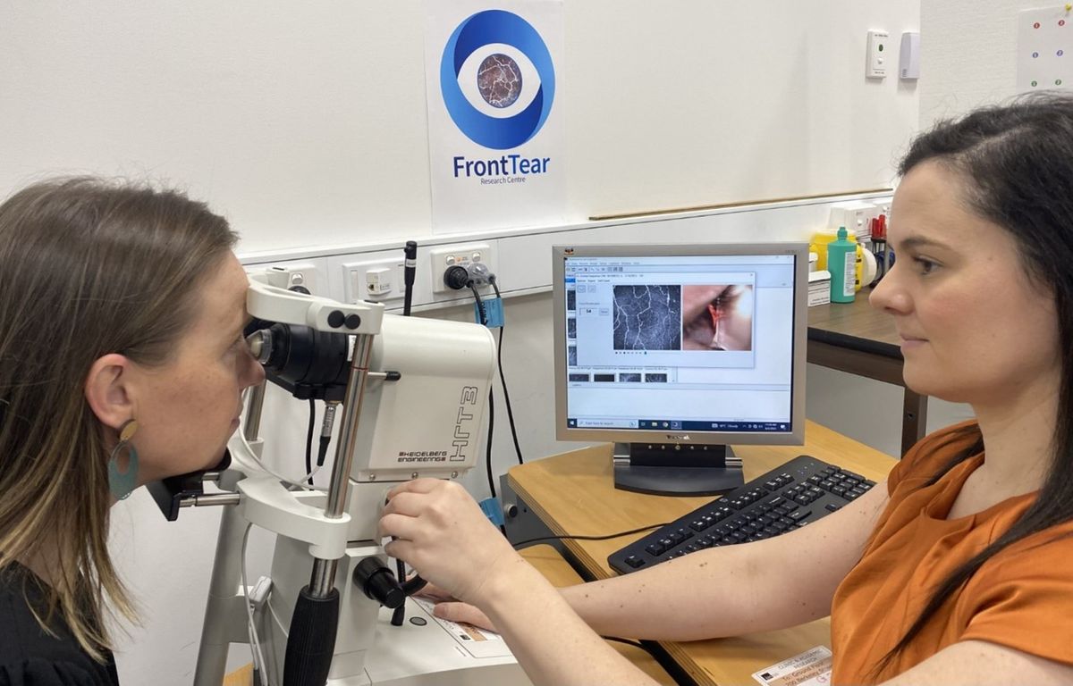 A woman sits in front of a confocal microscope to have her eyes checked, while an ocular immunologist adjusts the instrument. A computer on the desk behind them shows the images being captured.