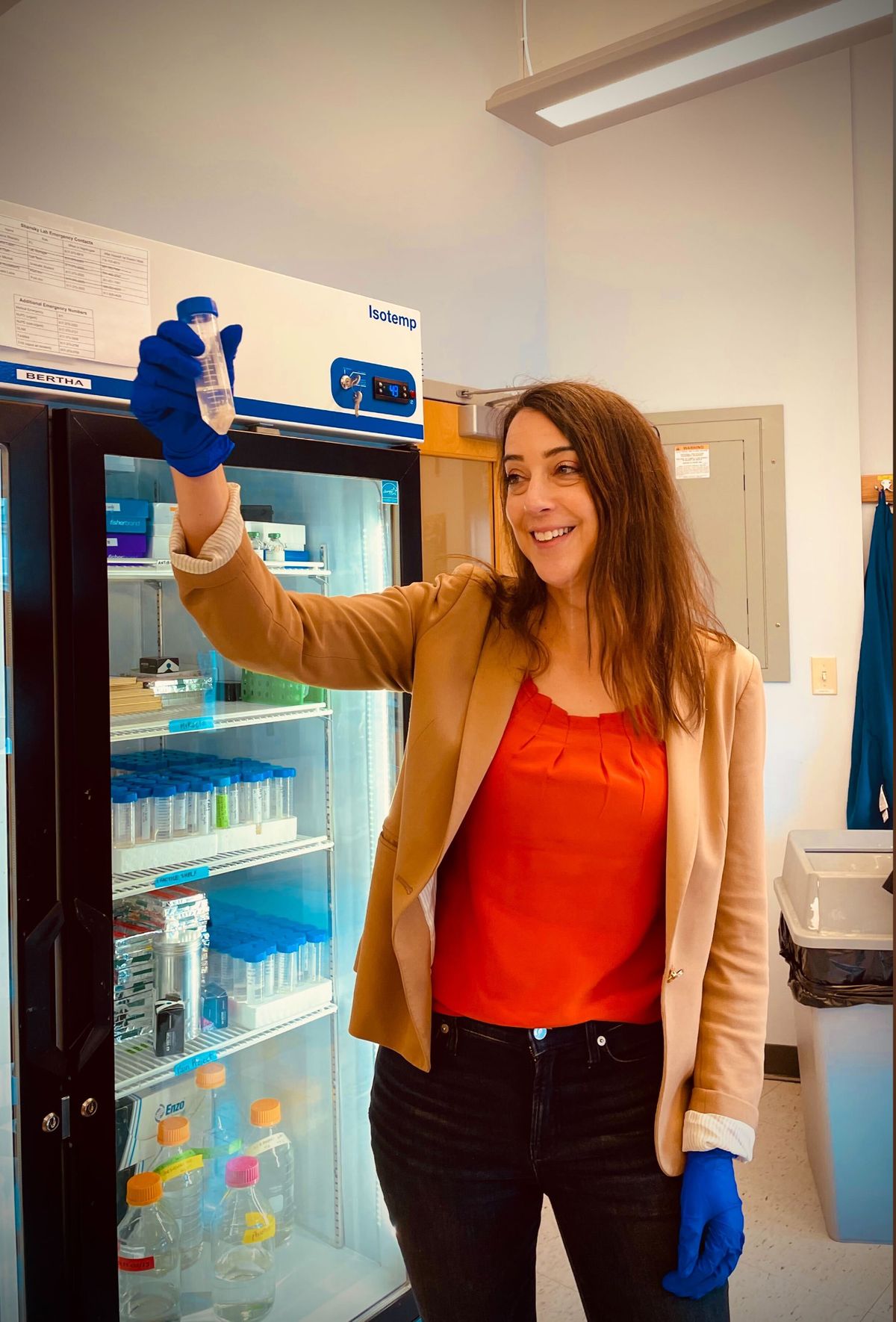 A female scientist is in a laboratory wearing blue gloves and holding a conical tube. She is standing in front of a refrigerator.