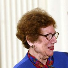 An older woman wearing a blue suit, shaking hands with an older man wearing a black suit.